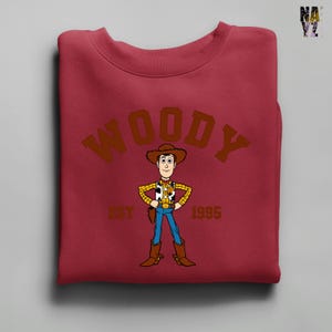 Puede incluir: Una sudadera granate con una imagen de dibujos animados de Woody de Toy Story. La palabra "WOODY" est&aacute; arqueada en la parte superior con letras marrones, con "EST 1995" debajo del personaje. Woody lleva sombrero de vaquero, chaleco, vaqueros azules y botas.