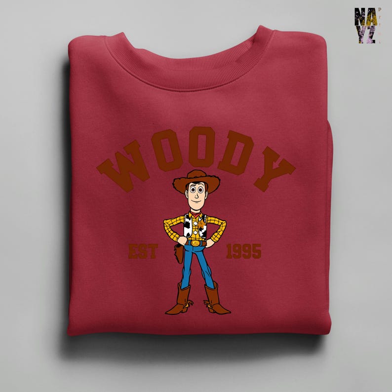Puede incluir: Una sudadera granate con una imagen de dibujos animados de Woody de Toy Story. La palabra "WOODY" est&aacute; arqueada en la parte superior con letras marrones, con "EST 1995" debajo del personaje. Woody lleva sombrero de vaquero, chaleco, vaqueros azules y botas.