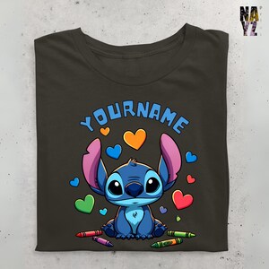 K&ouml;nnte beinhalten: Ein dunkelgraues T-Shirt mit einer blauen Stitch-Figur aus dem Disney-Film Lilo & Stitch. Stitch ist von bunten Herzen und Buntstiften umgeben. Der Text "YOURNAME" ist &uuml;ber Stitch gedruckt.