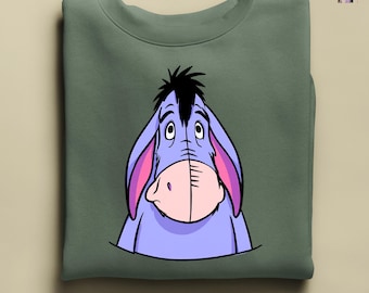 May include: An olive green sweatshirt featuring a large graphic of Eeyore from Winnie the Pooh. The cartoon donkey is depicted in shades of purple, pink, and black, with a sad expression. The sweatshirt is folded neatly.