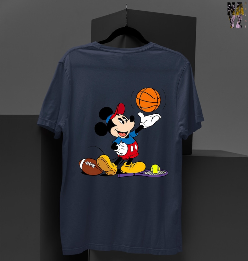 May include: Navy blue t-shirt featuring a cartoon Mickey Mouse character playing sports. Mickey is wearing a red hat, blue shirt, and red shorts. A basketball, football, and tennis racket with a ball are also visible.