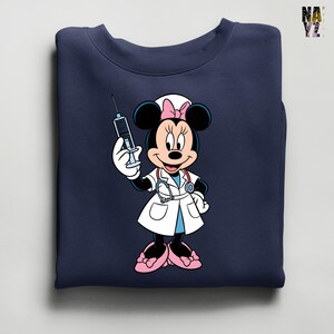 Peut inclure: Sweat-shirt bleu marine avec Minnie Mouse en infirmi&egrave;re tenant une seringue. Minnie porte une blouse blanche, des chaussures roses et un bonnet d'infirmi&egrave;re avec un n&oelig;ud rose. Le sweat-shirt est pli&eacute;.