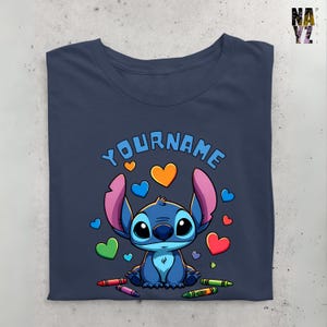 K&ouml;nnte beinhalten: Ein marineblaues T-Shirt mit einer Zeichentrickfigur Stitch, umgeben von bunten Herzen und Buntstiften. Der Text "YOUR NAME" ist &uuml;ber Stitch gedruckt.