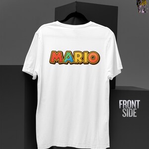 May include: White t-shirt with the word "MARIO" in colorful, cartoon-style lettering across the back. The text is outlined in brown with red, yellow, and blue coloring. The shirt is hanging on a black hanger.