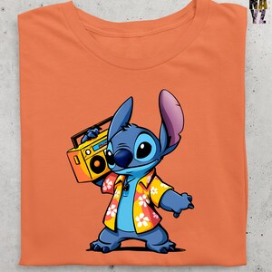 Op de afbeelding: Oranje T-shirt met een cartoon illustratie van Stitch uit Lilo & Stitch. Stitch is blauw met grote oren, draagt een Hawaiiaans shirt en houdt een gele boombox vast. Het T-shirt is netjes opgevouwen.