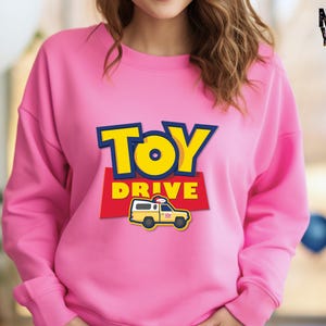 May include: Pink sweatshirt with a graphic of a yellow toy truck with the words "Toy Drive" in blue and red.