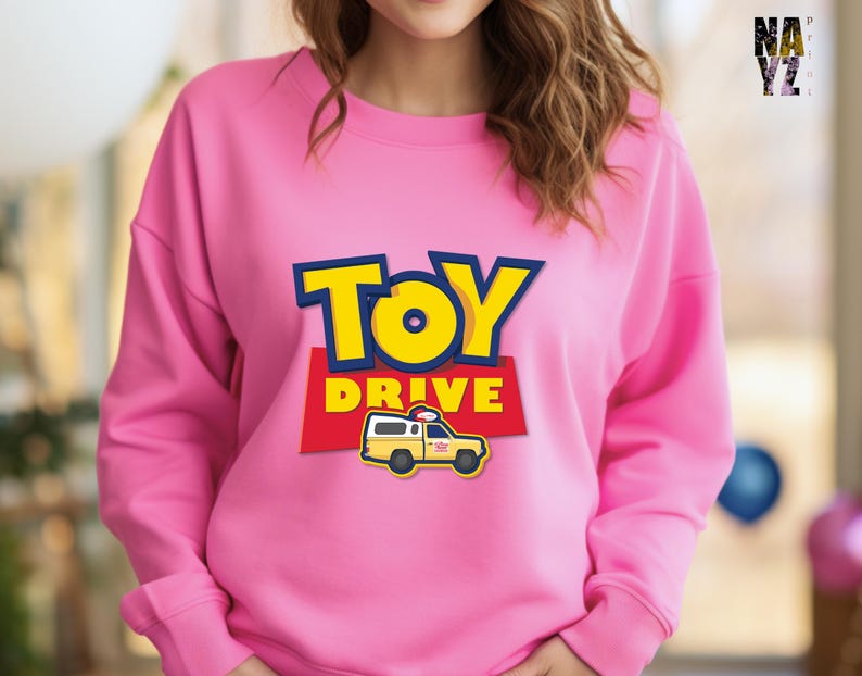 May include: Pink sweatshirt with a graphic of a yellow toy truck with the words "Toy Drive" in blue and red.