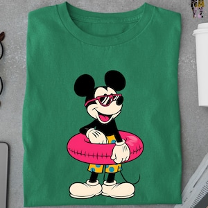 May include: Green t-shirt featuring a cartoon Mickey Mouse wearing sunglasses and holding a pink inflatable ring. Mickey is wearing yellow shorts with blue polka dots. The shirt is made of a soft material.