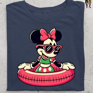 May include: Navy blue t-shirt with a cartoon Minnie Mouse design. Minnie Mouse is wearing sunglasses and a pink bow, and is in a red and pink inflatable ring. She is wearing a green and red polka dot dress. The shirt is folded.