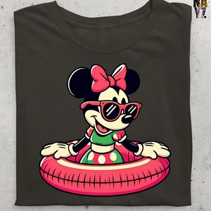 May include: Dark olive green t-shirt featuring a cartoon Minnie Mouse in a pink inner tube and sunglasses. Minnie wears a green and red polka dot dress and a pink bow. The shirt has a classic, cartoon style.