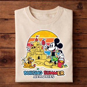 May include: Beige t-shirt featuring a colorful graphic of Mickey Mouse building a sandcastle with Pluto on a beach. The design includes a sunset backdrop and the text "BUILDING SUMMER MEMORIES".