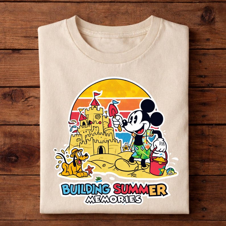 May include: Beige t-shirt featuring a colorful graphic of Mickey Mouse building a sandcastle with Pluto on a beach. The design includes a sunset backdrop and the text "BUILDING SUMMER MEMORIES".
