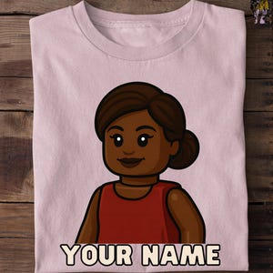 May include: A light pink t-shirt featuring a cartoon illustration of a person with dark skin, brown hair, and a red top. The text "YOUR NAME" is printed in a bold, black font at the bottom of the shirt.