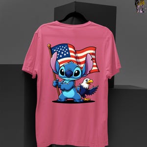 May include: Pink t-shirt featuring a cartoon Stitch character holding an American flag with an eagle. The design includes red, white, and blue colors. The shirt is a crew neck style.