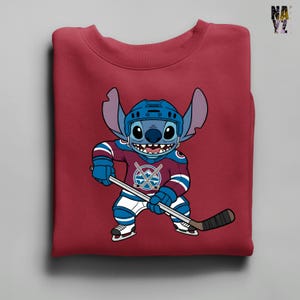 Puede incluir: Una sudadera granate con un personaje de dibujos animados, Stitch, vestido con equipo de hockey. Stitch lleva un casco azul y una camiseta granate y azul con un logotipo de palos de hockey cruzados. Sostiene un palo de hockey y lleva patines.