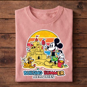 May include: A light pink t-shirt featuring a colorful graphic of Mickey Mouse building a sandcastle with Pluto. The design includes a sunset backdrop and the text "BUILDING SUMMER MEMORIES".