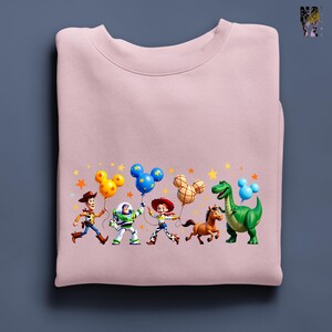 Puede incluir: Sudadera rosa claro con un gr&aacute;fico colorido de personajes de Toy Story. Los personajes sostienen globos con forma de Mickey Mouse sobre un fondo de estrellas.