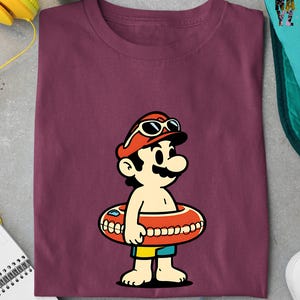 May include: Burgundy t-shirt featuring a cartoon character wearing a red hat, sunglasses, and an orange inner tube. The character has a large mustache and is wearing yellow and blue shorts. The design is centered on the shirt.