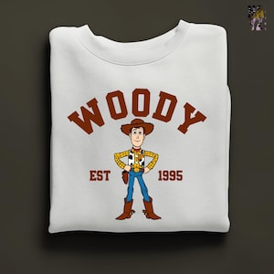 Puede incluir: Sudadera blanca con una ilustraci&oacute;n de dibujos animados de Woody de Toy Story. La palabra "WOODY" en marr&oacute;n est&aacute; arqueada en la parte superior, con "EST 1995" debajo. El dise&ntilde;o est&aacute; centrado en la prenda doblada, una prenda cl&aacute;sica.