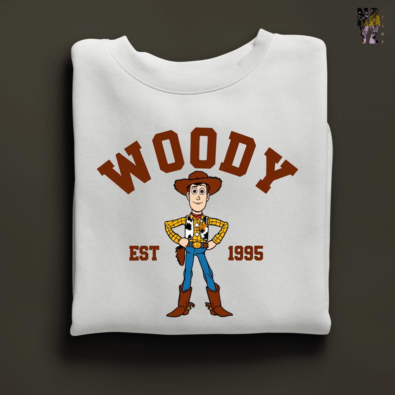 Puede incluir: Sudadera blanca con una ilustraci&oacute;n de dibujos animados de Woody de Toy Story. La palabra "WOODY" en marr&oacute;n est&aacute; arqueada en la parte superior, con "EST 1995" debajo. El dise&ntilde;o est&aacute; centrado en la prenda doblada, una prenda cl&aacute;sica.
