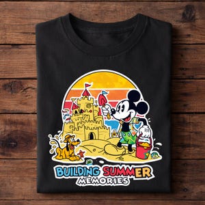 May include: Black t-shirt featuring a colorful graphic of Mickey Mouse building a sandcastle with Pluto at the beach. The design includes a sunset backdrop and the text "BUILDING SUMMER MEMORIES".
