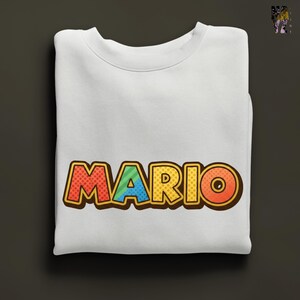 May include: White sweatshirt with the name "MARIO" in large, colorful, comic-style lettering. The letters are outlined in brown with red, blue, yellow, and orange fill. The sweatshirt is folded neatly on a dark gray surface.
