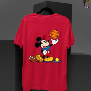 May include: Red t-shirt featuring a cartoon Mickey Mouse character. Mickey is depicted in a blue shirt, red shorts, and yellow shoes, holding a basketball. A football and tennis racket are also present.