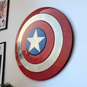 May include: A large, round shield with a red outer ring, a silver middle ring, and a blue center featuring a white star. The shield is mounted on a white wall, suggesting a decorative or collectible item.