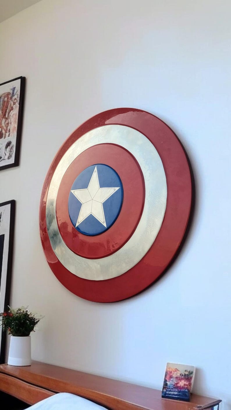 May include: A large, round shield with a red outer ring, a silver middle ring, and a blue center featuring a white star. The shield is mounted on a white wall, suggesting a decorative or collectible item.