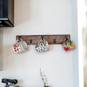 Op de afbeelding: Een houten wandbevestigde bekerrek met drie kleurrijke bekers die aan haken hangen. De bekers hebben bloem- en abstracte ontwerpen.