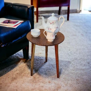 Pode incluir: Uma pequena mesa de madeira redonda com três pernas, com uma superfície marrom texturizada. Uma chaleira branca com desenhos florais, um jarro de creme branco e um açucareiro branco são colocados na mesa.