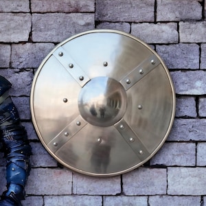 May include: A silver round shield with a raised center and four spokes extending from the center to the edge. The shield is mounted on a brick wall.