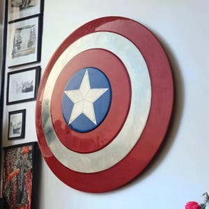 May include: A Captain America shield, a circular emblem with a red, white, and blue color scheme. The shield features a white star on a blue circle, surrounded by a silver ring and a red outer ring. It is mounted on a white wall.