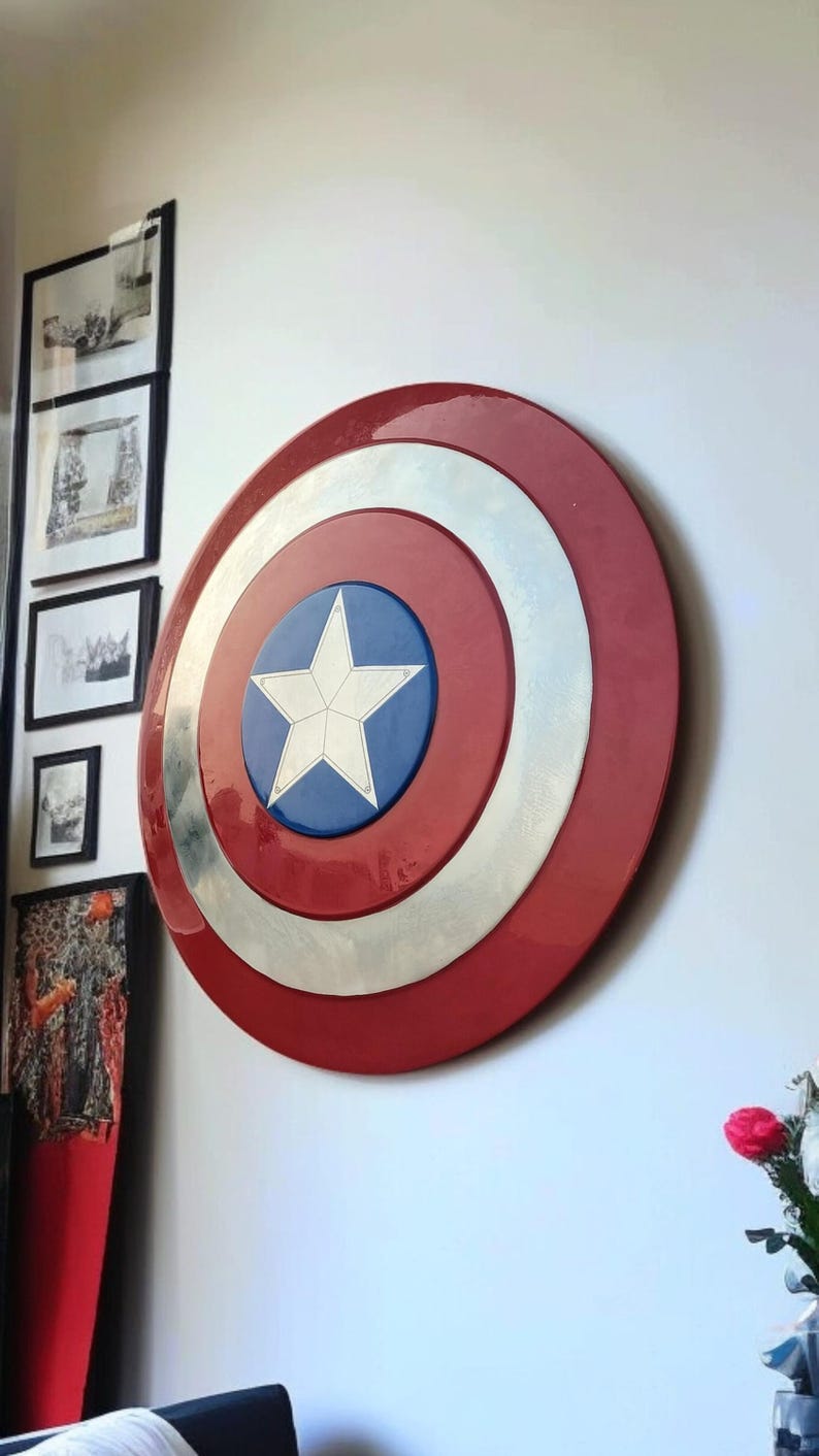 May include: A Captain America shield, a circular emblem with a red, white, and blue color scheme. The shield features a white star on a blue circle, surrounded by a silver ring and a red outer ring. It is mounted on a white wall.