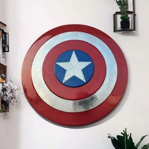 May include: A Captain America shield, a circular, red, white, and blue shield with a white star in the center. The shield is mounted on a white wall. The shield's diameter is approximately 30 inches.