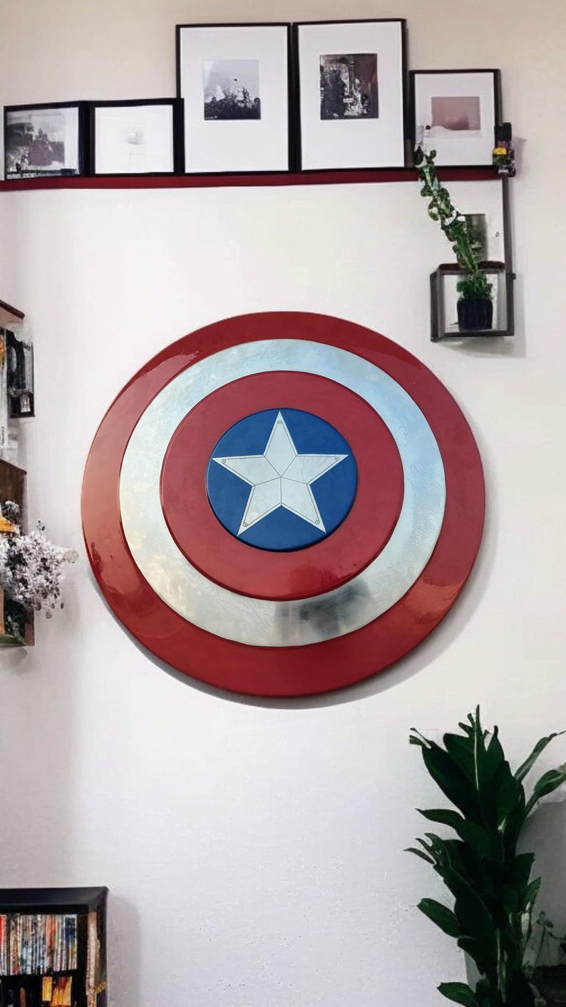 May include: A Captain America shield, a circular, red, white, and blue shield with a white star in the center. The shield is mounted on a white wall. The shield's diameter is approximately 30 inches.