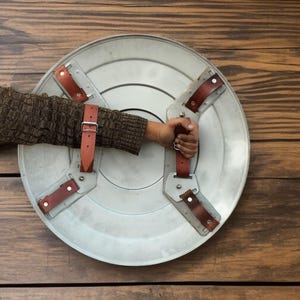 May include: A silver-colored shield with brown leather straps and handle. The shield is round with a slightly textured surface. The straps are riveted to the shield. The handle is held by a person.