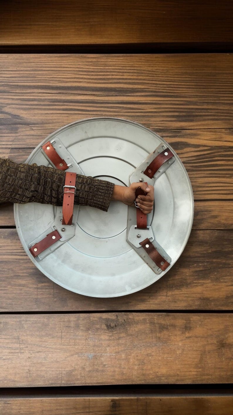 May include: A silver-colored shield with brown leather straps and handle. The shield is round with a slightly textured surface. The straps are riveted to the shield. The handle is held by a person.