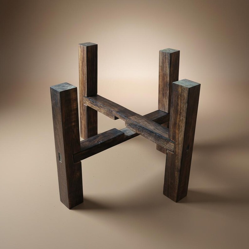 Puede incluir: Un soporte de madera marr&oacute;n oscuro para plantas con una base cuadrada y cuatro soportes verticales. El soporte tiene un dise&ntilde;o en forma de cruz en el centro, proporcionando una base estable para una planta en maceta. La madera tiene una veta natural.