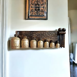 Puede incluir: Una estantería de pared de madera con intrincados tallados, que sostiene un conjunto de ocho campanas doradas de diferentes tamaños. La estantería también presenta dos clavijas de madera con remates decorativos.