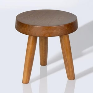May include: A small, round wooden stool with three legs. The stool has a natural wood finish, with a circular seat and angled legs. The wood grain is visible, and the stool appears to be handcrafted.