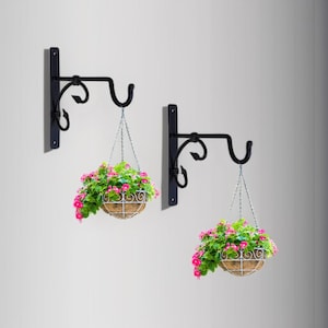 Op de afbeelding: Twee zwarte metalen hangende mandbeugels met decoratief krulwerk. Elke beugel ondersteunt een hangende mand gevuld met roze en groene bloemen. De manden hangen aan zilveren kettingen.