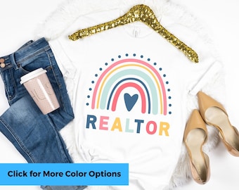 May include: White t-shirt with a rainbow and heart design and the word "REALTOR" printed on it. The shirt is on a hanger and there are jeans and a coffee cup in the background.