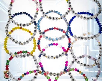 Può includere: Una collezione di braccialetti di perline con lettere bianche che compongono frasi come Kiss All The Time, Late Night Talking, Golden, Watermelon Sugar, Harry, As It Was, Pop, Adore You, Together e Disco, alternate a perline di vetro colorate.