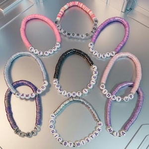Puede incluir: Ocho pulseras circulares hechas de pequeñas cuentas de arcilla planas y coloridas y cuentas con letras blancas. Las pulseras incluyen las frases WILD WORLD TOUR, EYE KONS, KATSEYE, GAME BOY, TONIGHT I MIGHT, PINKY UP, MY WAY e ICONIC BY MISTAKE.