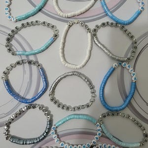 May include: A collection of twelve elastic bracelets made from small, flat, circular clay beads in shades of white, light blue, and silver. Each bracelet features white or silver letter beads spelling phrases like Backstreet Boys, Millennium, and Vegas Sphere.
