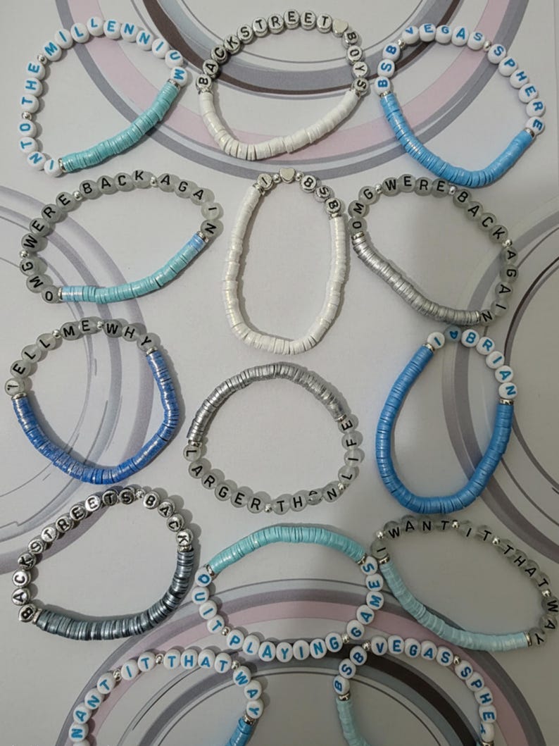 May include: A collection of twelve elastic bracelets made from small, flat, circular clay beads in shades of white, light blue, and silver. Each bracelet features white or silver letter beads spelling phrases like Backstreet Boys, Millennium, and Vegas Sphere.