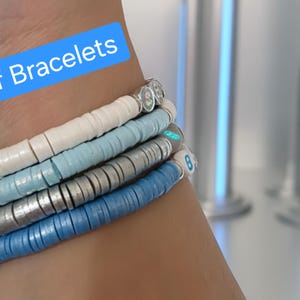 May include: A stack of four stretch bracelets made of flat, circular clay beads in white, light blue, silver, and medium blue. The bracelets feature small, round accent beads with letters and numbers. A blue text box reads Back of Bracelets.