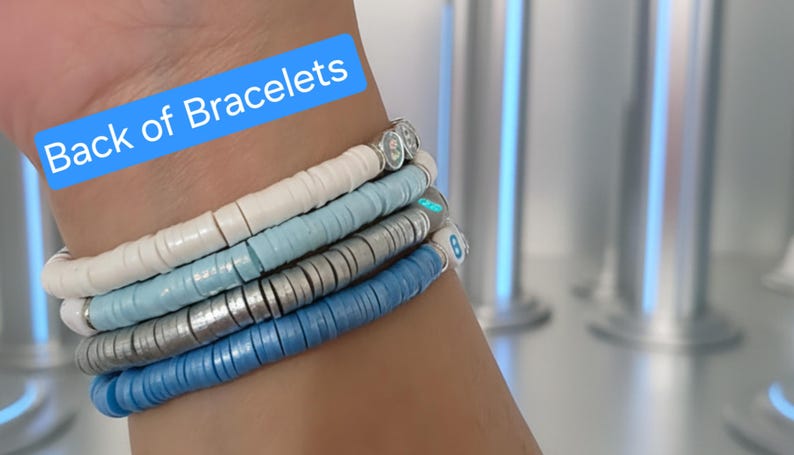 May include: A stack of four stretch bracelets made of flat, circular clay beads in white, light blue, silver, and medium blue. The bracelets feature small, round accent beads with letters and numbers. A blue text box reads Back of Bracelets.