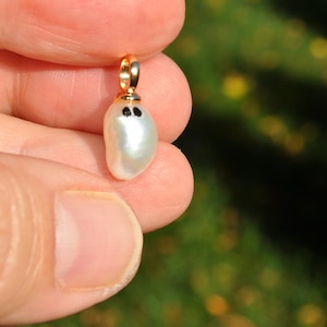 May include: A small, pear-shaped pearl pendant with a gold bail. The pearl has two small black dots, giving it a whimsical appearance. The pendant is held between fingers against a blurred green background.
